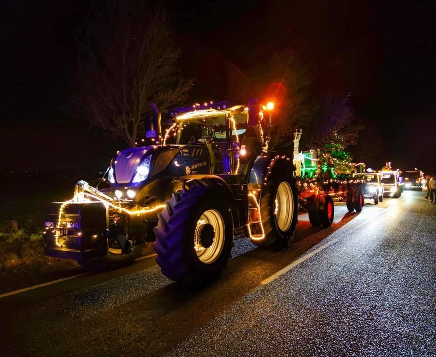 NM- Nerchau-Mutzschener - Landwirtschaft - Ein blauer, mit Lichterketten geschmückter Traktor führt einen nächtlichen Fahrzeugumzug durch eine Straße.