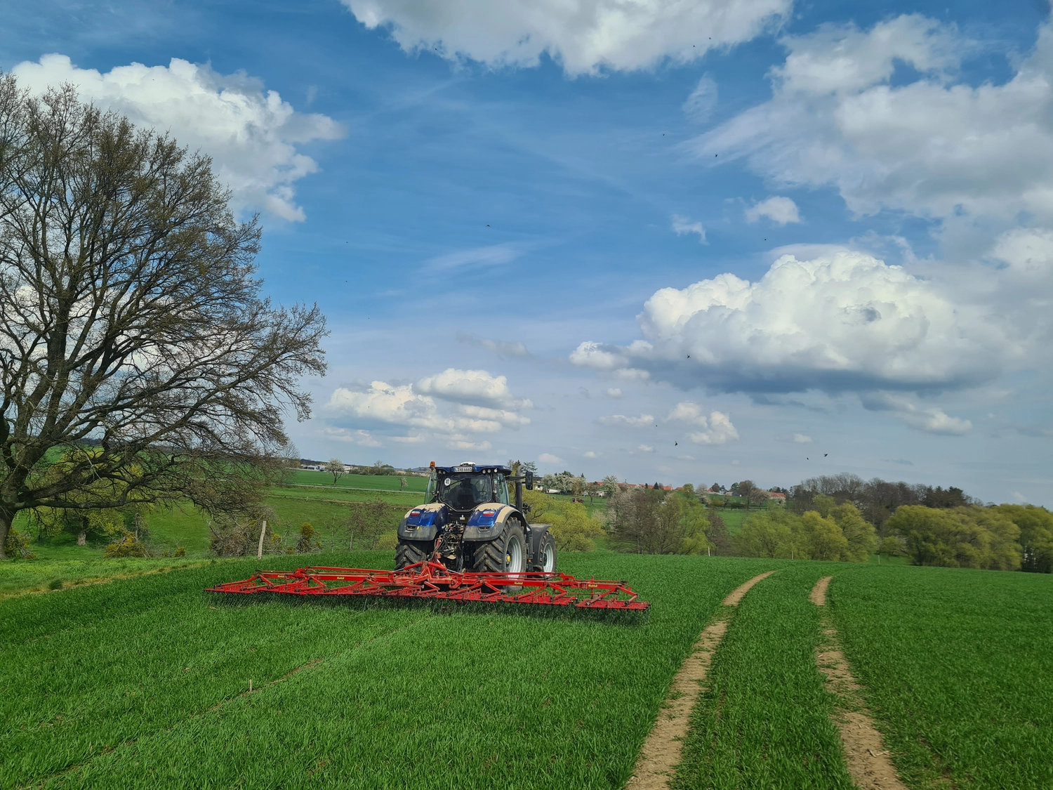 NM- Nerchau-Mutzschener - Landwirtschaft - Ein Traktor zieht ein breites, rotes Unkrautbekämpfungsgerät durch ein grünes Feld unter bewölktem Himmel.